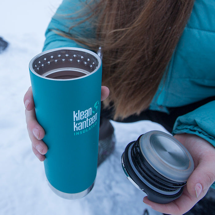 Woman crouched in snow holding Klean Kanteen TKWide insulated mug in one hand and lid in the other